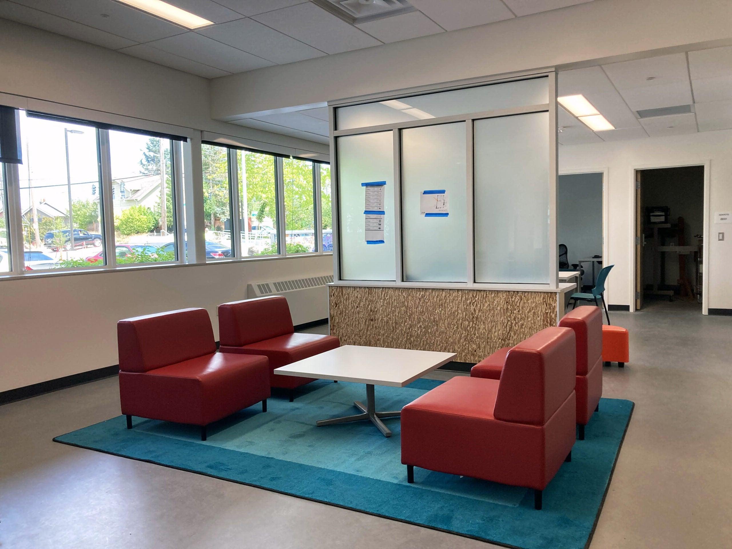 four red chairs sit on a blue area rug with a table between them. a partial wall separates them from another area. windows are on one wall