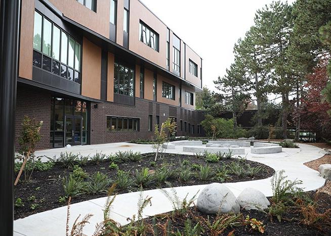 a paved walkway leads to a patio area that has seating. a three story building is behind it