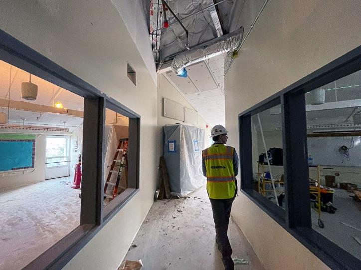 a person stands in a mostly complete hallway with windows on each side. the ceiling has ductwork showing and the floor is protected.